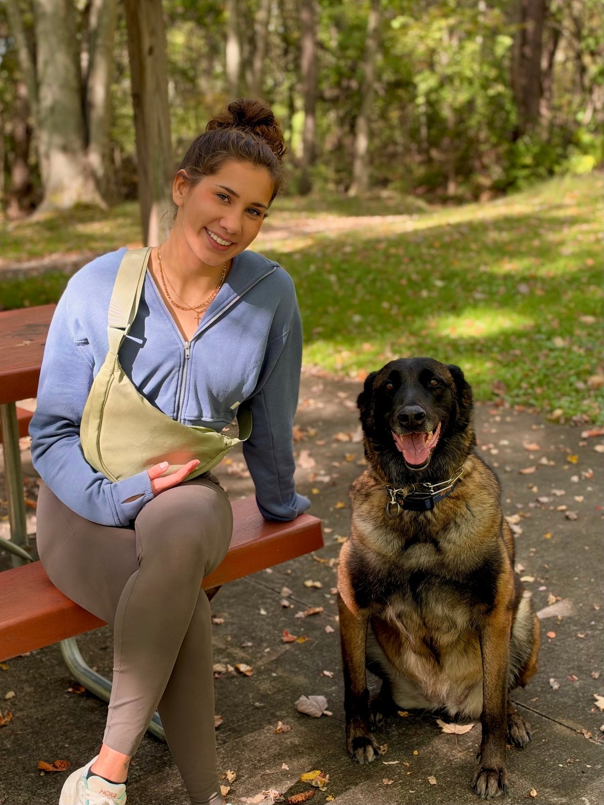Elena smiling on a park picnic table next to Mishka, her Belgian Shepherd, sitting and smiling at the camera
